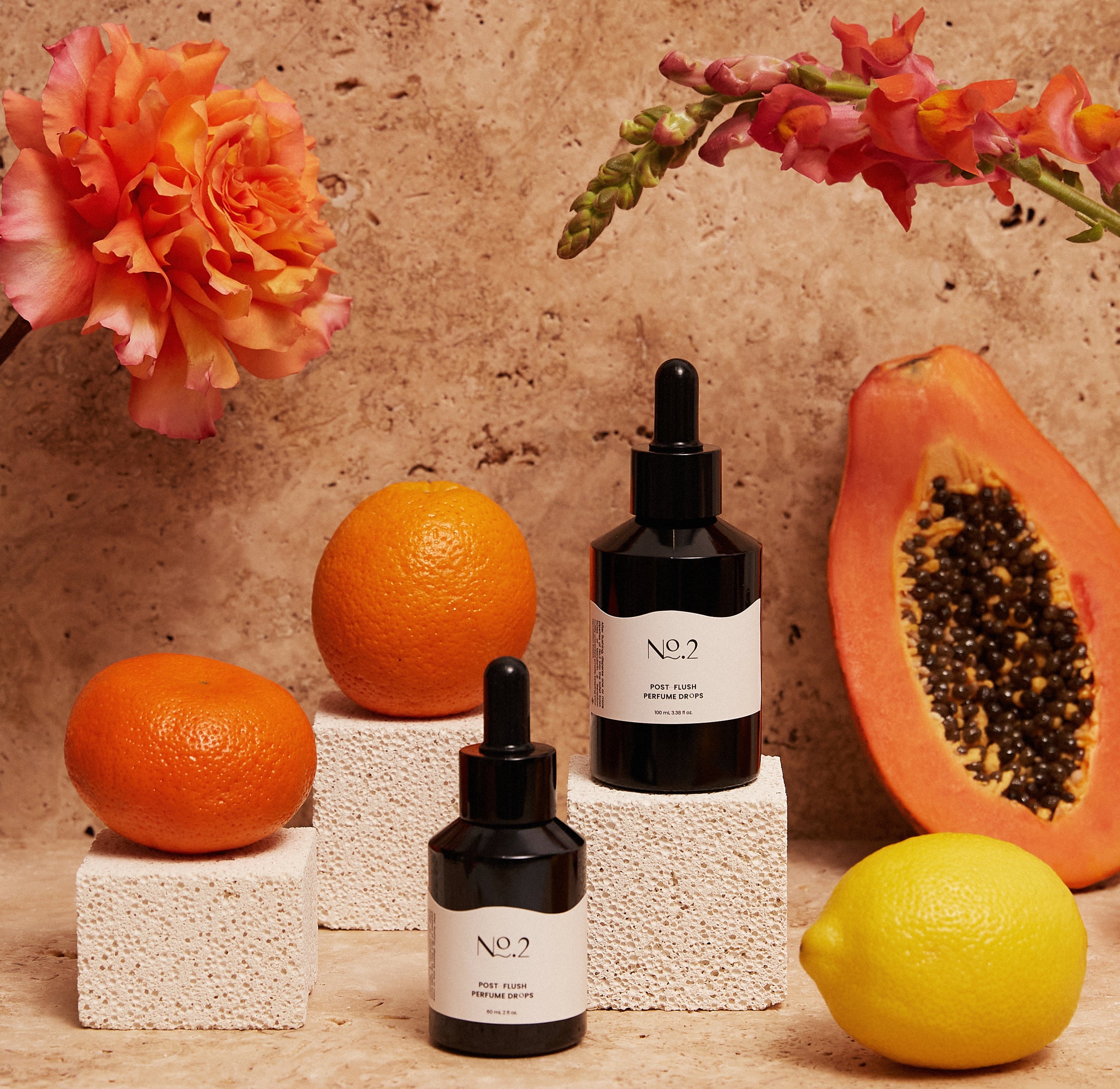 Two bottles of No.2 Co post poo toilet drops, surrounded by a mandarin, lemon and an orange. Two orange flowers and a sand coloured tile in the background